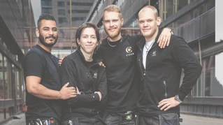 Fire smilende kollegaer i arbeidsklær med Coor-logoen foran en bygning.