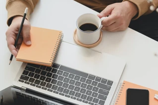 Laptop sett ovenifra med hender som holder notatblokk og kaffe